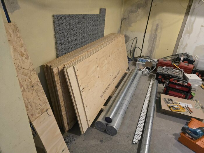 Plywood och OSB-skivor lutande mot en vägg, omgivna av byggmaterial som ventilationsrör, verktyg och paneler i ett förråd under renovering.