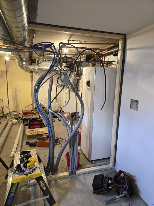 Byggarbetsplats med ventilationsrör, elslangar och verktyg; värmepump i bakgrunden, pågående installation.