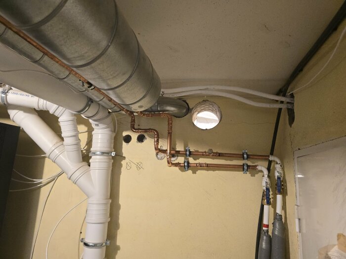 Ventilationssystem i ett rum med olika rör för frånluft och tilluft, inklusive kopparrör och plastslangar, installerade på en gul vägg.