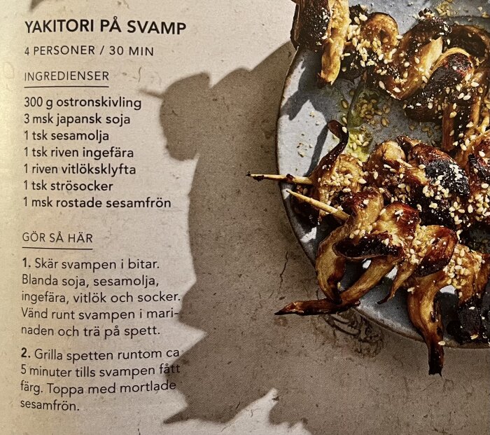 Grillade yakitori-spett med svamp liggande på ett fat, garnerade med rostade sesamfrön. Recept och ingredienser synliga.