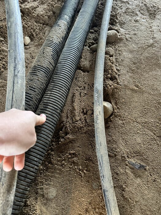 Hand pekar på svart korrugerat rör i sand, rör kopplas till bergvärmepump i garage, diskussion om byte och extra skyddsrör innan gjutning.