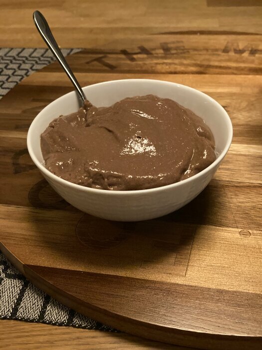 Vit skål med chokladpudding och en sked på ett träbord.