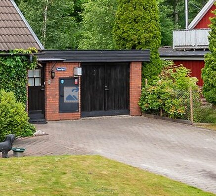 70-tals garage med tjärpapptak, svart dubbelport och tegelväggar, omgiven av trädgård. Planerar att bygga carport i samma stil med plåttak.