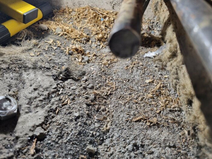 Närbild av ett lutande elementrör ovanför sågspån och isolering, med ett gult verktyg i bakgrunden.