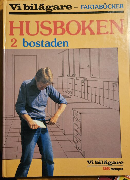 Man på bokomslag undersöker ett golvplan i ett kök; boken heter "Husboken 2: Bostaden" från Vi Bilägare faktaböcker.