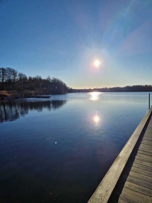 Solig dag vid en sjö med en brygga som sträcker sig ut i vattnet, omgiven av träd i bakgrunden, med solens reflektion synlig på ytan.