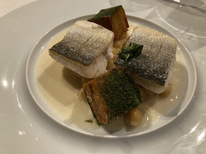 Två bitar vit fisk med skinn serverade med brödbitar och krämig sås på en vit tallrik.