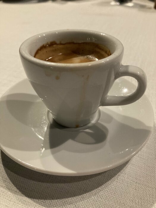 Vit kaffekopp med espresso på ett vitt fat på en duk.