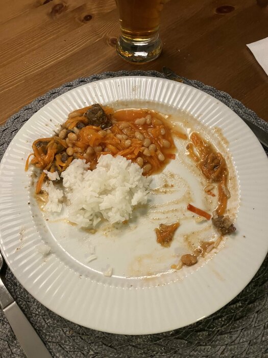 Tallrik med rester av chiligryta med vita bönor och rivna morötter, en portion ris bredvid. Glas med dryck i bakgrunden på ett träbord.