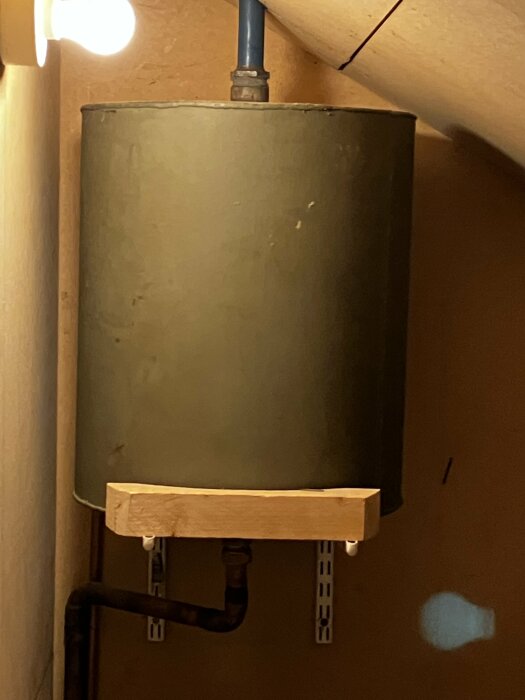 Bild på en cylindrisk, metallisk tank i ett trångt utrymme, monterad med röranslutningar och trästöd. Fråga om den kan tas bort.