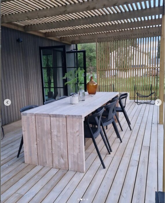 Pergola med stående ribbor som insynsskydd, lång träbord med svarta stolar på trädäck, omgiven av byggnad med liggande panel och fönster.