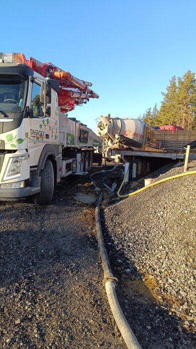 Betongpumpbil och cementblandare på arbetsplats med slangar på marken för markgjutning, omgivna av grus och trä.