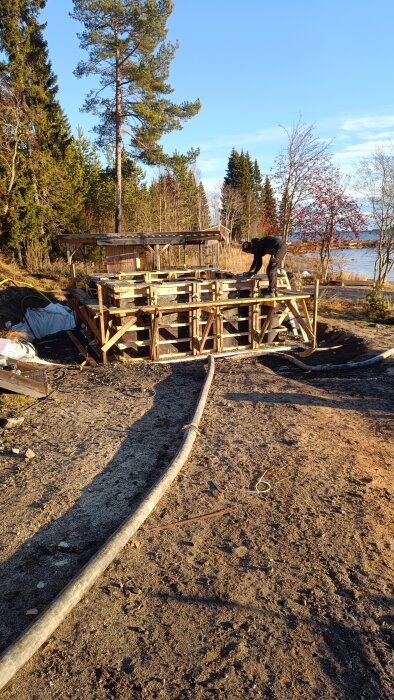 Person bygger form för betonggjutning med hjälp av markslang i träbeklädd ställning; omgivande skog och sjö i bakgrunden.