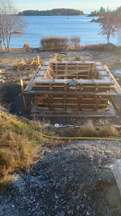 Byggform för gjutning vid strandkant med sjöutsikt och trädridå i bakgrunden.