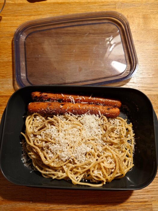 Korvonaran-matlåda med spaghetti, riven ost och korv i en svart plastlåda på träbord.