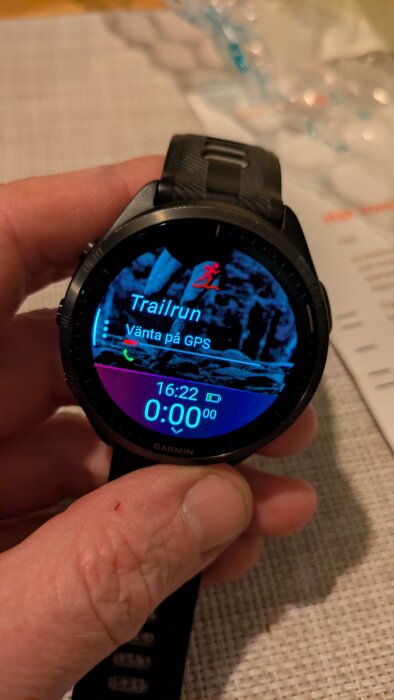 En hand håller en Garmin Fenix 6X-klocka, skärmen visar "Trailrun" och "Vänta på GPS" med tid och batteristatus.