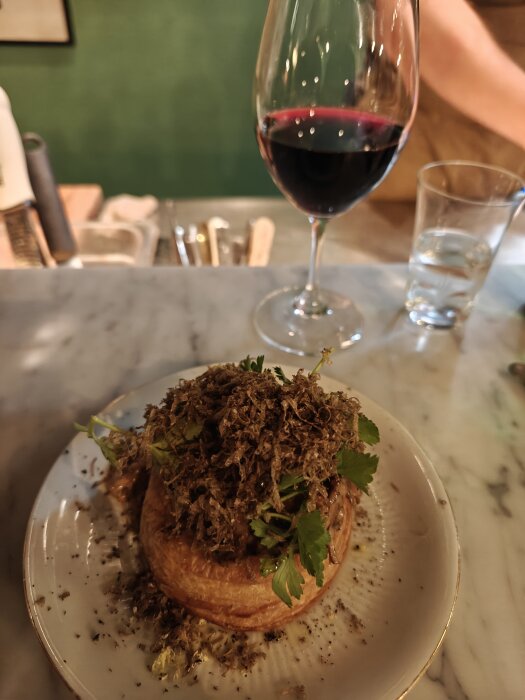 Bakverk toppat med örter och torkad svamp på fat och ett glas rödvin på ett marmorbord.