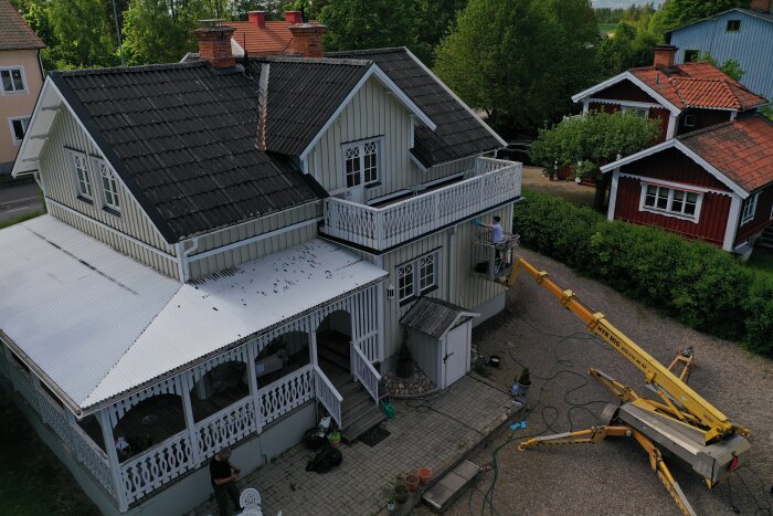 Luftfoto av ett äldre hus med vitmålad veranda och plasttak. En person målar fasaden från en lyftkran; omgivningen inkluderar träd och närområdesbyggnader.