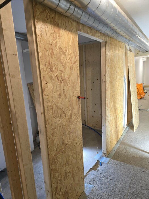 OSB-skivor i inre konstruktion med ventilationsrör i taket, synlig öppning mot duschrum under renovering. Byggmaterial och delvis färdigt golv.