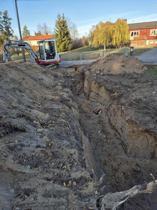 Grävningsarbete för dränering och VA-byte vid ett 50-talshus, grävmaskin synlig, sandig mark och grus, hus och träd i bakgrunden.