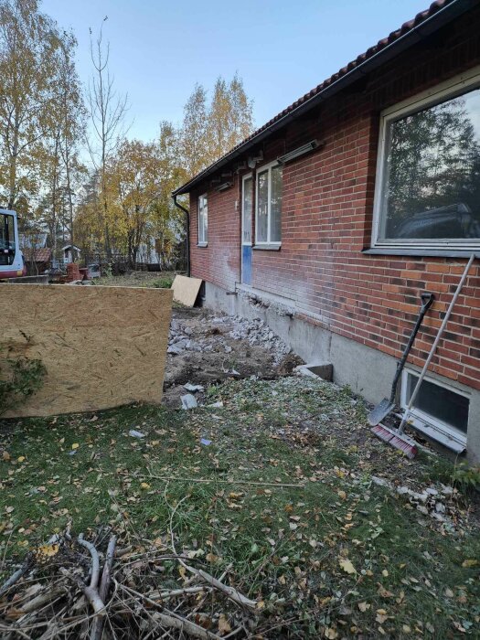 Pågående dränering vid tegelhus, betongplatta och jord bortgrävd. Verktyg vid husvägg, höstlöv och grenar på gräsmattan.