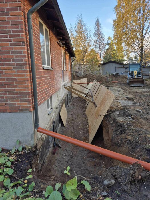 Dräneringsarbete vid ett tegelhus med öppet dike och träram för stöd, orange avloppsrör synligt. Grävmaskin i bakgrunden.