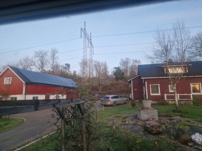 Röd byggnad med solpaneler på taket, grusväg, bil och trädgård med sten och krukor i förgrunden, elledningar i bakgrunden.