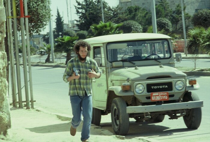 Man går bredvid en beige Toyota Land Cruiser i Amman, 1982, med kamerautrustning i handen.