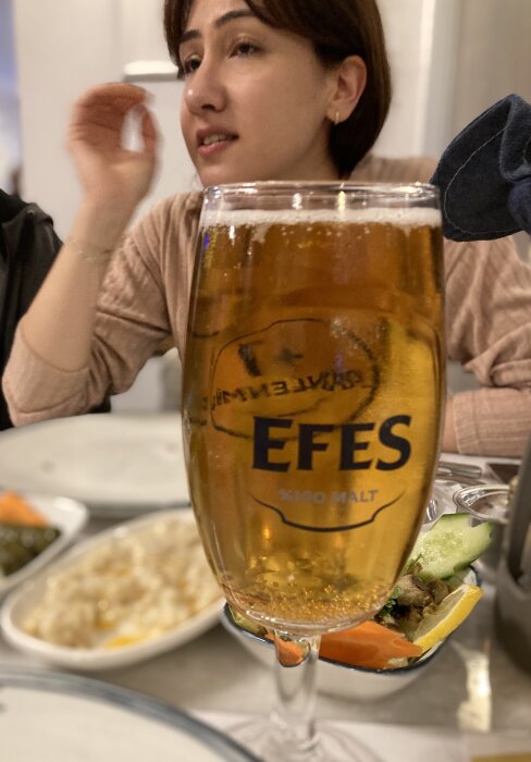 Ett glas öl märkt Efes på ett bord med mat och en person i bakgrunden.