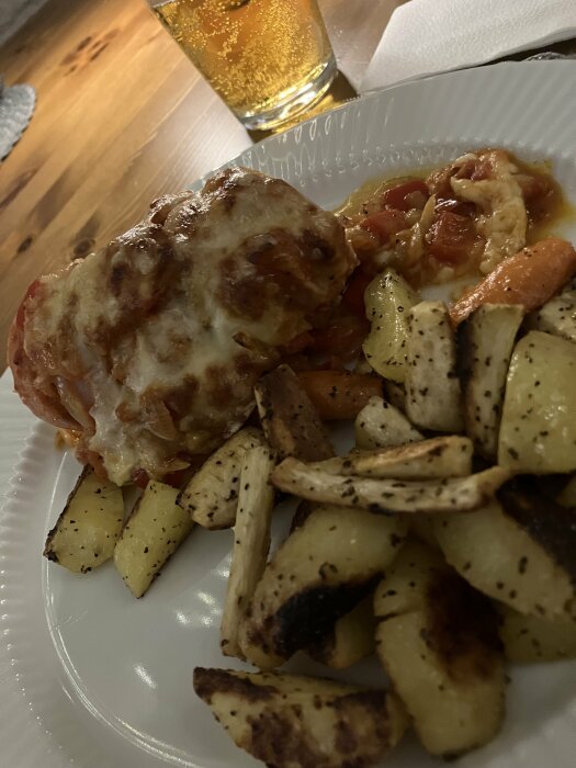 Tallrik med falukorv i ugn med smält ost och tomatsås samt ugnsrostade rotfrukter. Glas med dryck i bakgrunden.