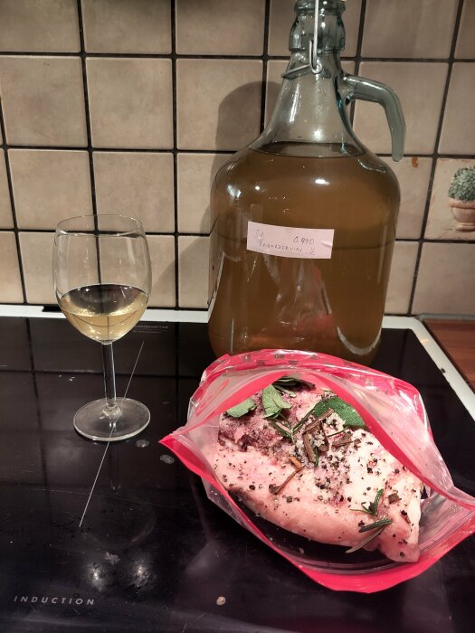 En stor flaska vitt vin, ett vinglas och en påse med kryddad guanciale på en spishäll.