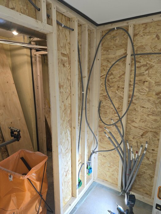 Väggkonstruktion med träramar och OSB-skiva, flera el- och nätverksrör installerade, orange sopsäck och bygglampa synliga.