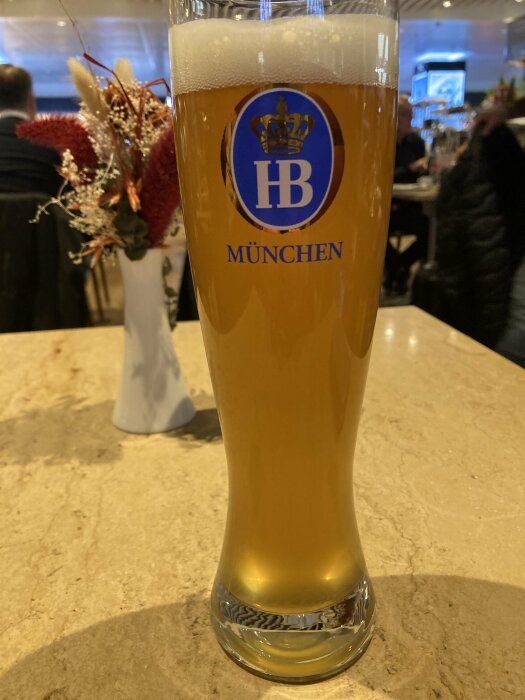Ett högt ölglas med HB-logotypen och texten "München", fyllt med gyllene öl på ett bord i restaurangmiljö, med vas och torkade blommor i bakgrunden.