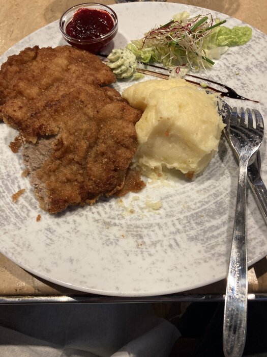 Panerad schnitzel med potatismos, serverad med lingonsylt, sallad och örtsmör på en tallrik.