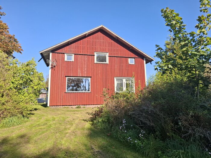 Rött trähus från 1800-talet omgiven av grönska mot en klarblå himmel.