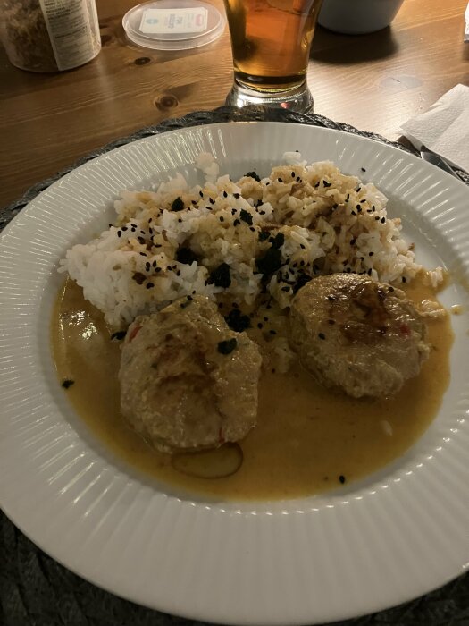 Thaibiffar i röd currysås med kokosmjölk och ris på tallrik, sesamfrön och kryddor ovanpå, bredvid ett glas dryck på ett matbord.