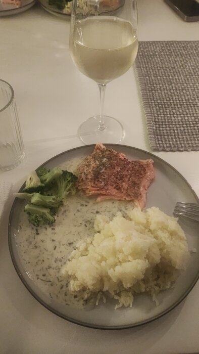 Tallrik med lax, pressad potatis, Sandefjordssås och broccoli, bredvid ett glas vitt vin på ett dukat bord.