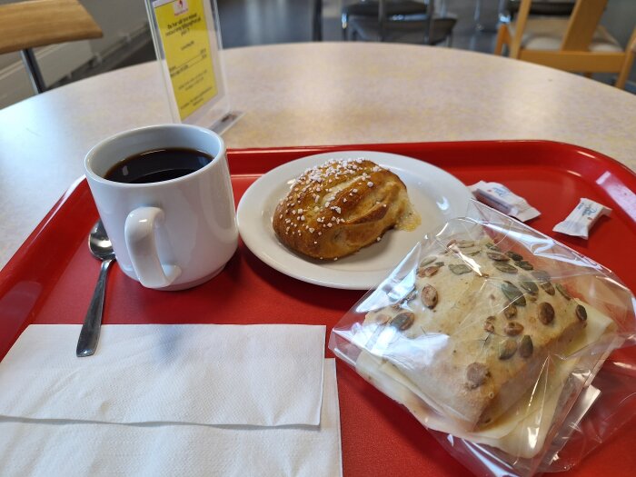 Kaffe och bulle på vit tallrik, fralla i plastpåse med pumpafrön, servett och sked på röd bricka på ett bord.