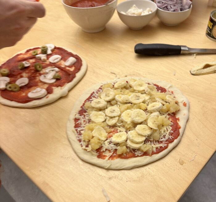 Två odekorerade pizzor på ett träbord; en med bananer och ost, den andra med tomatsås, svamp och oliver. Skålar med pizzapålägg i bakgrunden.