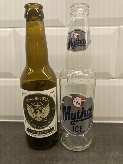 Två tomma ölflaskor, en från Fenix Brewery Double Bock och en Mythos Ice, står på en grå yta framför vitt kakel.