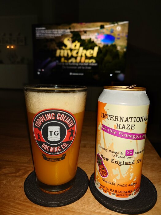 Glas fylld med New England IPA från Toppling Goliath, med en burk av märket International Haze i bakgrunden, TV:n visar "Så mycket bättre".