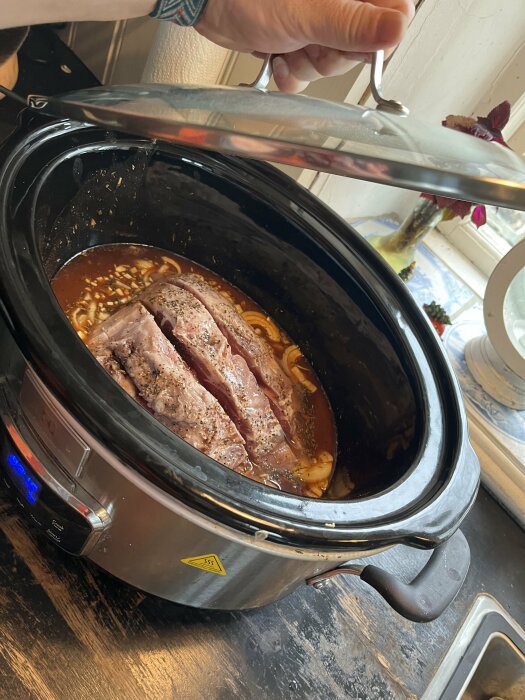 Pullad fläskkött tillagas i en slow cooker på en köksbänk, omgiven av en hand som lyfter locket.