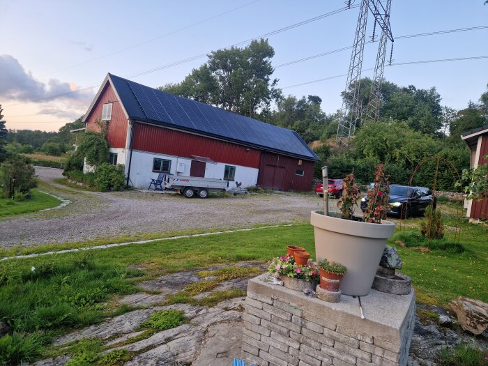 Röd ladugård med solpaneler på taket, grusväg framför, omgiven av grönska och trädgård med krukväxter i förgrunden.