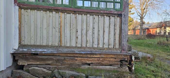 Sliten nederdel av punschveranda med omålad träpanel och rutten trästomme ovanpå en grund av stenblock.