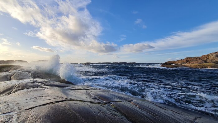 Havsvågor slår mot klipporna vid Tjurpannan i Bohuslän under en solig dag med ljusa moln på himlen.