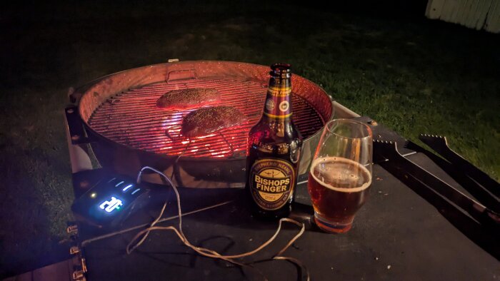 Flaska Bishop's Finger öl och ett glas öl bredvid en grill med köttbitar på natten.