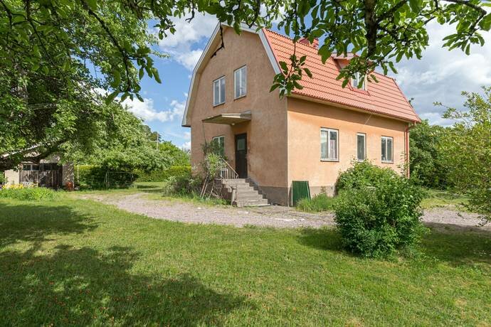 Gammalt hus från 1940-talet med tegelrött tak och putsad fasad, omgiven av grönskande trädgård och grusgång, redo för renovering.