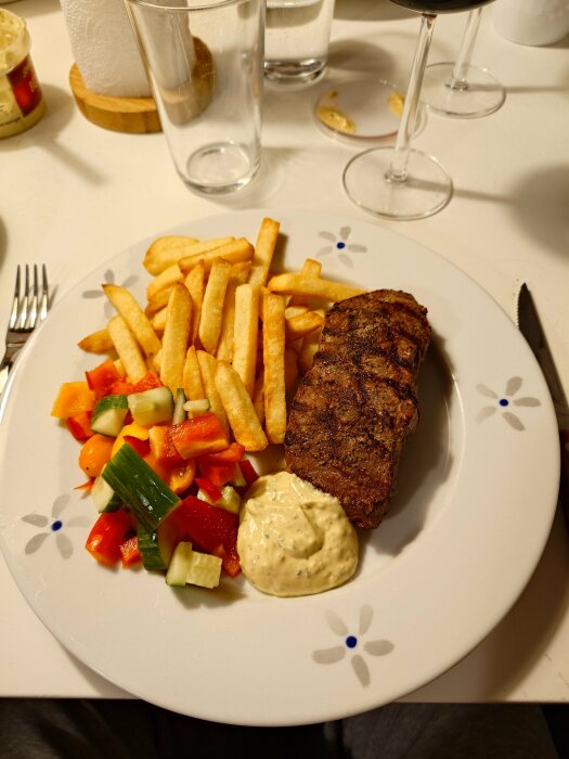 Tallrik med grillad biff, pommes frites, grönsaksbitar och sås. Background: glas och vinglas.