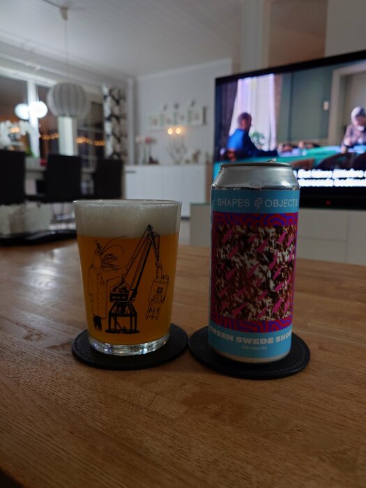 En burk "Shapes & Objects - Green Swede Shoes" DDH hazy IPA och ett glas öl står på ett träbord i ett inrett rum med en tv i bakgrunden.