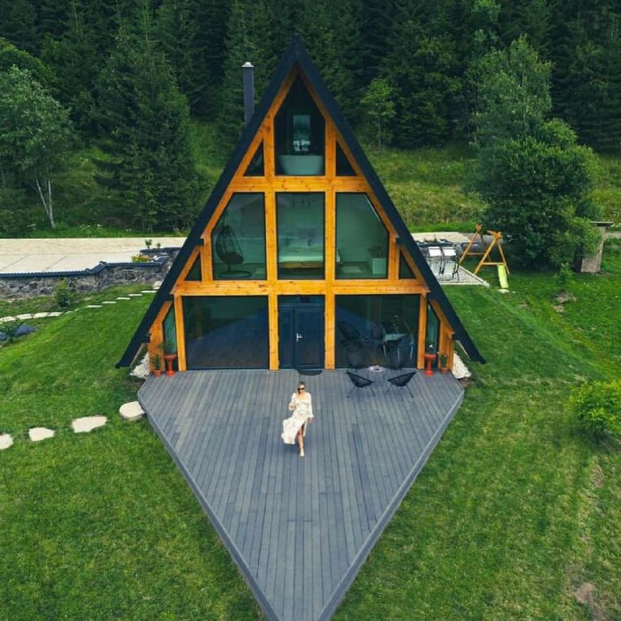 A-frame house with 60° roof, large windows, wooden terrace, greenery and a person walking on the deck.